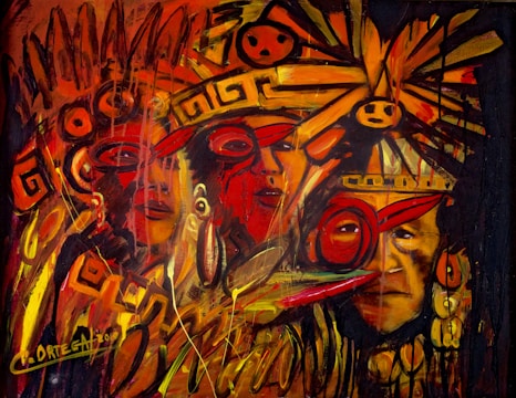 A vivid and energetic painting featuring abstract indigenous faces adorned with vibrant headgear and ornaments. The composition is filled with swirling patterns and symbols, predominantly in warm shades of red, orange, and yellow. The brushstrokes are dynamic, suggesting movement and intensity.