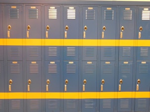 A row of identical blue lockers with a horizontal yellow stripe across the middle. Each locker features a vented door, chrome-colored handle, and lock. The lockers are neatly arranged in a school or gym setting.
