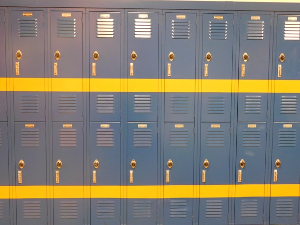 A row of identical blue lockers with a horizontal yellow stripe across the middle. Each locker features a vented door, chrome-colored handle, and lock. The lockers are neatly arranged in a school or gym setting.