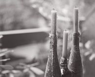 Three tall candles are placed in the necks of decorative bottles, with wax dripped down the sides. The scene is captured in a soft, blurred background, suggesting a serene and tranquil setting.