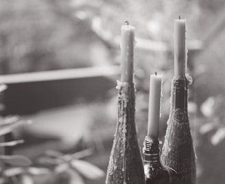 Three tall candles are placed in the necks of decorative bottles, with wax dripped down the sides. The scene is captured in a soft, blurred background, suggesting a serene and tranquil setting.