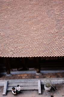 A vast array of traditional roof tiles extends across the image, creating a textured pattern. Below, two people are standing on a stone-paved area in front of the building, engaged in conversation. A large stone planter with a small plant is positioned nearby. The architecture and materials suggest a traditional setting.