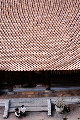 A vast array of traditional roof tiles extends across the image, creating a textured pattern. Below, two people are standing on a stone-paved area in front of the building, engaged in conversation. A large stone planter with a small plant is positioned nearby. The architecture and materials suggest a traditional setting.