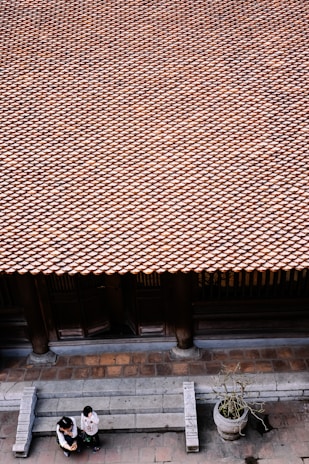 A vast array of traditional roof tiles extends across the image, creating a textured pattern. Below, two people are standing on a stone-paved area in front of the building, engaged in conversation. A large stone planter with a small plant is positioned nearby. The architecture and materials suggest a traditional setting.