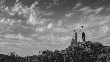 A couple holding hands stands atop a rocky cliff against a dramatic sky filled with scattered clouds, conveying a sense of adventure and romance.