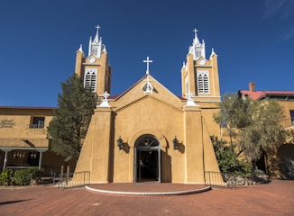 An adobe-style church with two bell towers and a tall central cross rises against a vivid blue sky. The structure features light tan walls, ornate white trim, and several black lanterns flanking the entrance. The surrounding area includes desert landscaping with shrubs and cacti.