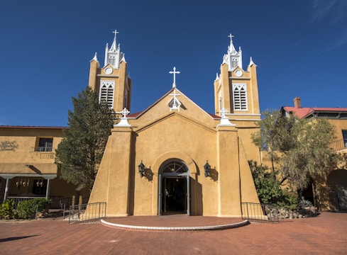An adobe-style church with two bell towers and a tall central cross rises against a vivid blue sky. The structure features light tan walls, ornate white trim, and several black lanterns flanking the entrance. The surrounding area includes desert landscaping with shrubs and cacti.