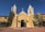 An adobe-style church with two bell towers and a tall central cross rises against a vivid blue sky. The structure features light tan walls, ornate white trim, and several black lanterns flanking the entrance. The surrounding area includes desert landscaping with shrubs and cacti.