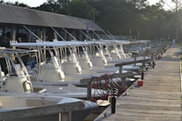 A row of white motorboats docked at a wooden pier under the roof of a marina, with trees in the background and various equipment such as red hoses and blue fenders visible.
