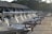 A row of white motorboats docked at a wooden pier under the roof of a marina, with trees in the background and various equipment such as red hoses and blue fenders visible.