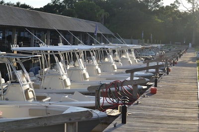 A row of white motorboats docked at a wooden pier under the roof of a marina, with trees in the background and various equipment such as red hoses and blue fenders visible.