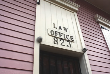 A close-up view of an exterior building wall with horizontal paneling painted in a soft pink color. The central focus is a large rectangular plaque stating 'LAW OFFICE 823' in bold letters. The paneling and design suggest a historical or vintage style.