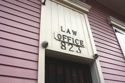 A close-up view of an exterior building wall with horizontal paneling painted in a soft pink color. The central focus is a large rectangular plaque stating 'LAW OFFICE 823' in bold letters. The paneling and design suggest a historical or vintage style.