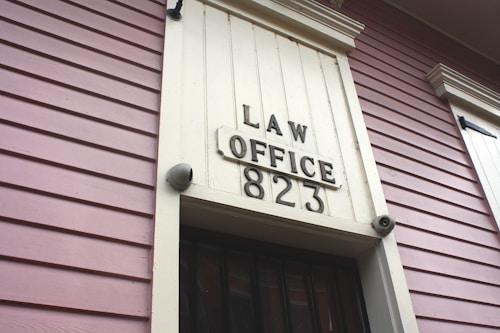 A close-up view of an exterior building wall with horizontal paneling painted in a soft pink color. The central focus is a large rectangular plaque stating 'LAW OFFICE 823' in bold letters. The paneling and design suggest a historical or vintage style.