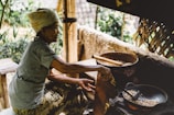 An elderly person is seated and roasting coffee beans over an open fire in a rustic outdoor setting. The person is wearing a headscarf and casual clothes. Near them, there is a wooden stool and a traditional woven bamboo structure in the background. The ambiance suggests a homely, rural environment.