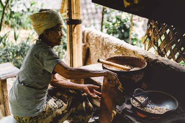 An elderly person is seated and roasting coffee beans over an open fire in a rustic outdoor setting. The person is wearing a headscarf and casual clothes. Near them, there is a wooden stool and a traditional woven bamboo structure in the background. The ambiance suggests a homely, rural environment.