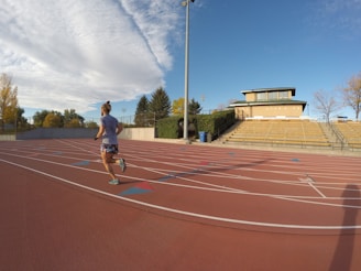 A person is jogging on an outdoor track with red running lanes. The track is surrounded by a stadium with empty yellow bleachers and a building in the background. The sky is clear and partially covered with a large, fluffy cloud, and there are trees with autumn leaves surrounding the area.
