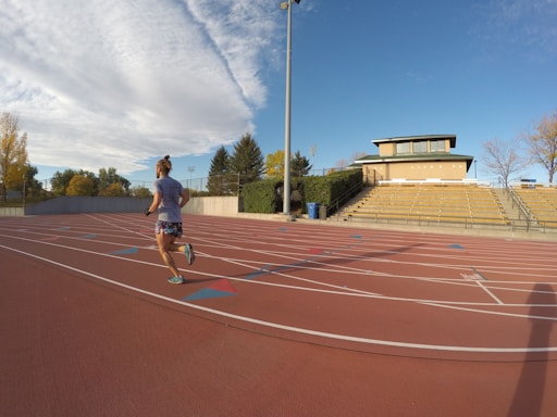 A person is jogging on an outdoor track with red running lanes. The track is surrounded by a stadium with empty yellow bleachers and a building in the background. The sky is clear and partially covered with a large, fluffy cloud, and there are trees with autumn leaves surrounding the area.