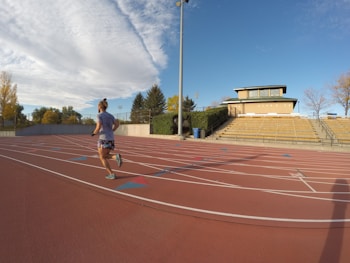 A person is jogging on an outdoor track with red running lanes. The track is surrounded by a stadium with empty yellow bleachers and a building in the background. The sky is clear and partially covered with a large, fluffy cloud, and there are trees with autumn leaves surrounding the area.