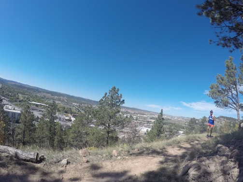 A person in athletic gear is running along a dirt trail surrounded by a forested area with tall pine trees. In the background, there is an expansive view of a town or city and a wide open sky with few clouds. The foreground features some rocks and a fallen tree trunk.