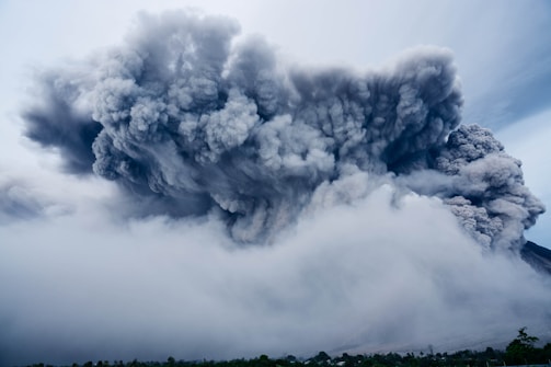 A massive cloud of ash and smoke billows into the sky from an erupting volcano. The thick, gray plumes dominate the image, with smaller clouds surrounding the larger cloud mass. The ground is obscured by a layer of haze and ash, suggesting a recent or ongoing eruption.