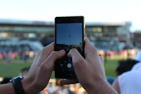 A person outdoors using an iPhone to photograph a sports event with Apex Zoom active.