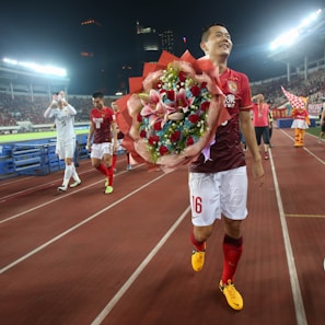 A football player in a red jersey smiles while holding a large bouquet of flowers. Other players and a mascot are visible in a stadium with bright lights and a cheering crowd in the background.