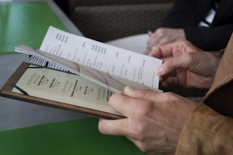 A person is holding a menu with both hands, flipping through the pages, which contain printed text. The setting appears to be a restaurant or cafe. Another person is seated nearby with their hands visible in the background.