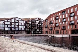 Two modern apartment buildings are situated alongside a waterfront. The building on the left features a dark facade with large windows, while the building on the right has a brick exterior. The foreground consists of a cobblestone walkway next to the water, creating a serene urban environment.