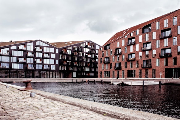 Two modern apartment buildings are situated alongside a waterfront. The building on the left features a dark facade with large windows, while the building on the right has a brick exterior. The foreground consists of a cobblestone walkway next to the water, creating a serene urban environment.