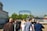 A group of people is walking towards a metal gate with a blue sign in Cyrillic script that reads 'Добро пожаловать' over it. The scene is set outdoors with greenery around and a bright sky visible. People are casually dressed, and some are carrying backpacks.