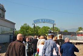 A group of people is walking towards a metal gate with a blue sign in Cyrillic script that reads 'Добро пожаловать' over it. The scene is set outdoors with greenery around and a bright sky visible. People are casually dressed, and some are carrying backpacks.