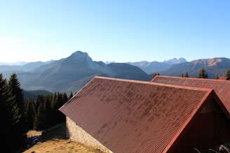 A rustic building with a red, corrugated metal roof is set against a backdrop of a mountain range. The peaks of the mountains are surrounded by a clear blue sky. Dense evergreen trees line the landscape in front of the mountains.