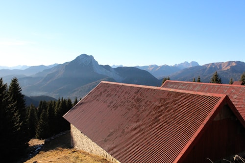 A rustic building with a red, corrugated metal roof is set against a backdrop of a mountain range. The peaks of the mountains are surrounded by a clear blue sky. Dense evergreen trees line the landscape in front of the mountains.