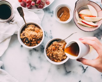 A breakfast scene featuring two bowls of oatmeal topped with granola. One hand pours syrup over one of the bowls. Nearby, there are plates of red grapes and apple slices, a bowl of cinnamon, and a cup of coffee. The setting is atop a marble surface with a white cloth in the background.