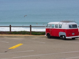 A red and white vintage van is parked near a scenic beach. The ocean can be seen in the background, along with several seagulls flying above the water. A wooden fence separates the parking area from the shore, and the sky appears overcast, giving a calm and serene atmosphere.