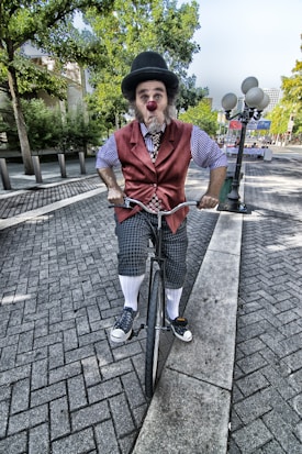 A clown with a red nose and hat is riding a unicycle along a paved path. He wears a red vest, checkered shorts, and high socks. Trees and a cityscape are visible in the background.