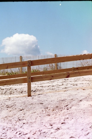 A sandy area with a simple wooden fence in the foreground, set against a clear blue sky with a few fluffy white clouds. The fence is composed of horizontal wooden planks supported by vertical posts, with sparse vegetation visible beyond.