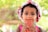 A young child wearing a white hat with red cherry patterns and a pink sleeveless outfit is holding a red rectangular object near their mouth. The background is softly blurred, with bright green foliage indicating outdoor lighting and a sunny day.