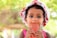 A young child wearing a white hat with red cherry patterns and a pink sleeveless outfit is holding a red rectangular object near their mouth. The background is softly blurred, with bright green foliage indicating outdoor lighting and a sunny day.