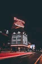 A historic building illuminated by a prominent neon sign that reads 'Dispatch Ohio's Greatest Home Newspaper'. The building is surrounded by city streets with blurred red and white lights from passing vehicles, indicating motion. Flags are visible on poles near the building, and a large advertisement is displayed on an adjacent building.
