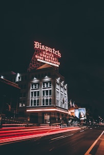 A historic building illuminated by a prominent neon sign that reads 'Dispatch Ohio's Greatest Home Newspaper'. The building is surrounded by city streets with blurred red and white lights from passing vehicles, indicating motion. Flags are visible on poles near the building, and a large advertisement is displayed on an adjacent building.