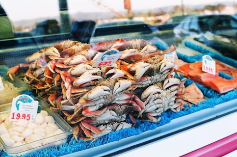 A seafood display features a pile of fresh crabs with visible claws and shells. Price tags are present, indicating cost per pound. Surrounding the crabs, there are packages containing scallops and other fish products. The display is lined with a blue mat, creating a visually appealing arrangement.