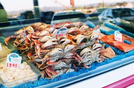 A seafood display features a pile of fresh crabs with visible claws and shells. Price tags are present, indicating cost per pound. Surrounding the crabs, there are packages containing scallops and other fish products. The display is lined with a blue mat, creating a visually appealing arrangement.
