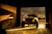 A vintage car without doors and front wheels is parked in a dimly lit garage, with sunlight streaming through the open door, casting shadows on the brick walls and concrete floor.
