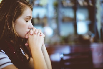 A young woman is seen with her hands clasped together near her face, possibly in a praying or thoughtful position. She has closed eyes and a calm expression. The background is blurred, indicating she is inside a dimly-lit room with shelves and various items in the background.