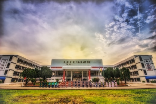 A wide-angle view of a large, multi-story school building with a central entrance and flags displayed. A group of people is lined up in front of the entrance, and there are trees and a well-maintained lawn in the foreground. The sky above is partially cloudy, creating a dramatic backdrop.