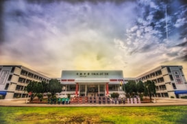A wide-angle view of a large, multi-story school building with a central entrance and flags displayed. A group of people is lined up in front of the entrance, and there are trees and a well-maintained lawn in the foreground. The sky above is partially cloudy, creating a dramatic backdrop.