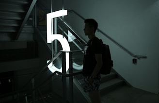 A person stands next to a large illuminated number '5' in a stairwell, casting a silhouette. The setting is dimly lit, with shadows creating a moody atmosphere. The stair railings and steps are visible, contributing to the architectural elements of the scene.