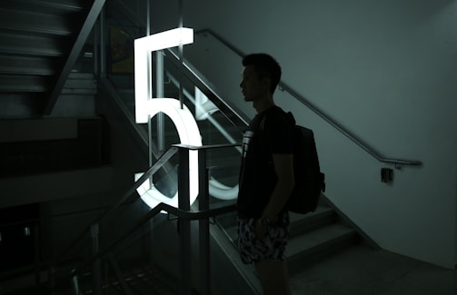 A person stands next to a large illuminated number '5' in a stairwell, casting a silhouette. The setting is dimly lit, with shadows creating a moody atmosphere. The stair railings and steps are visible, contributing to the architectural elements of the scene.
